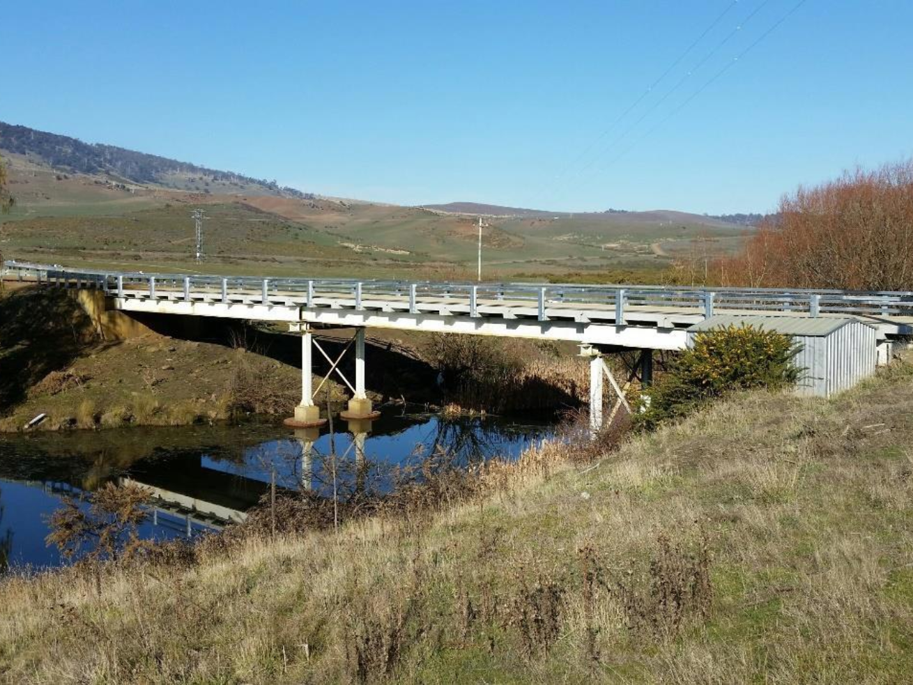 The Rotherwood Bridge Re-Deck features a metal bridge with guardrails spanning over a calm river in a rural landscape, surrounded by grassy hills and distant mountains under a clear blue sky.