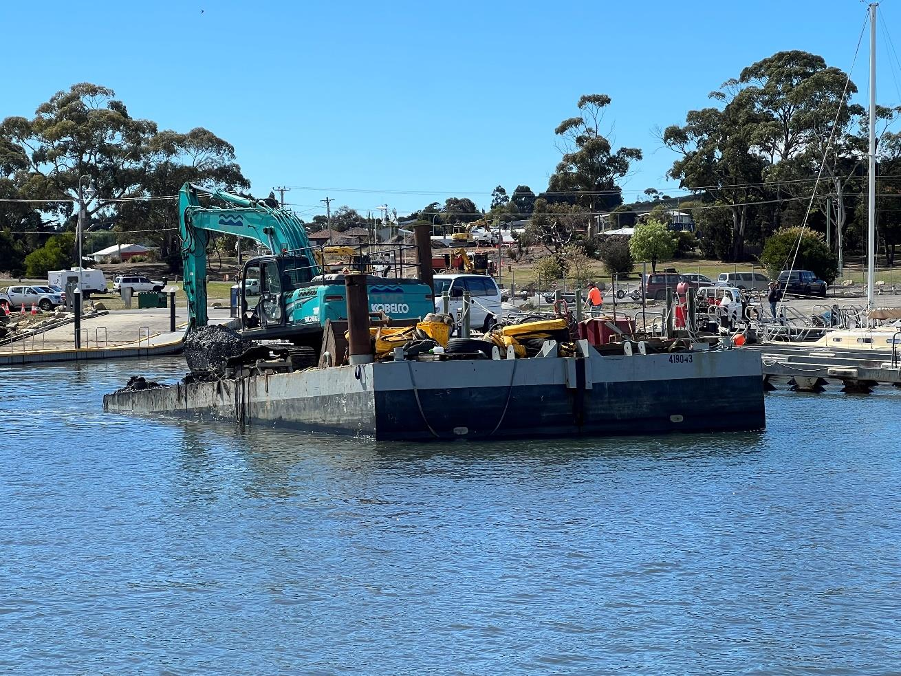 Excavators dredging the marina approach channel, removing a mixture of mud, sand, and rock, with stockpiled materials visible onshore.